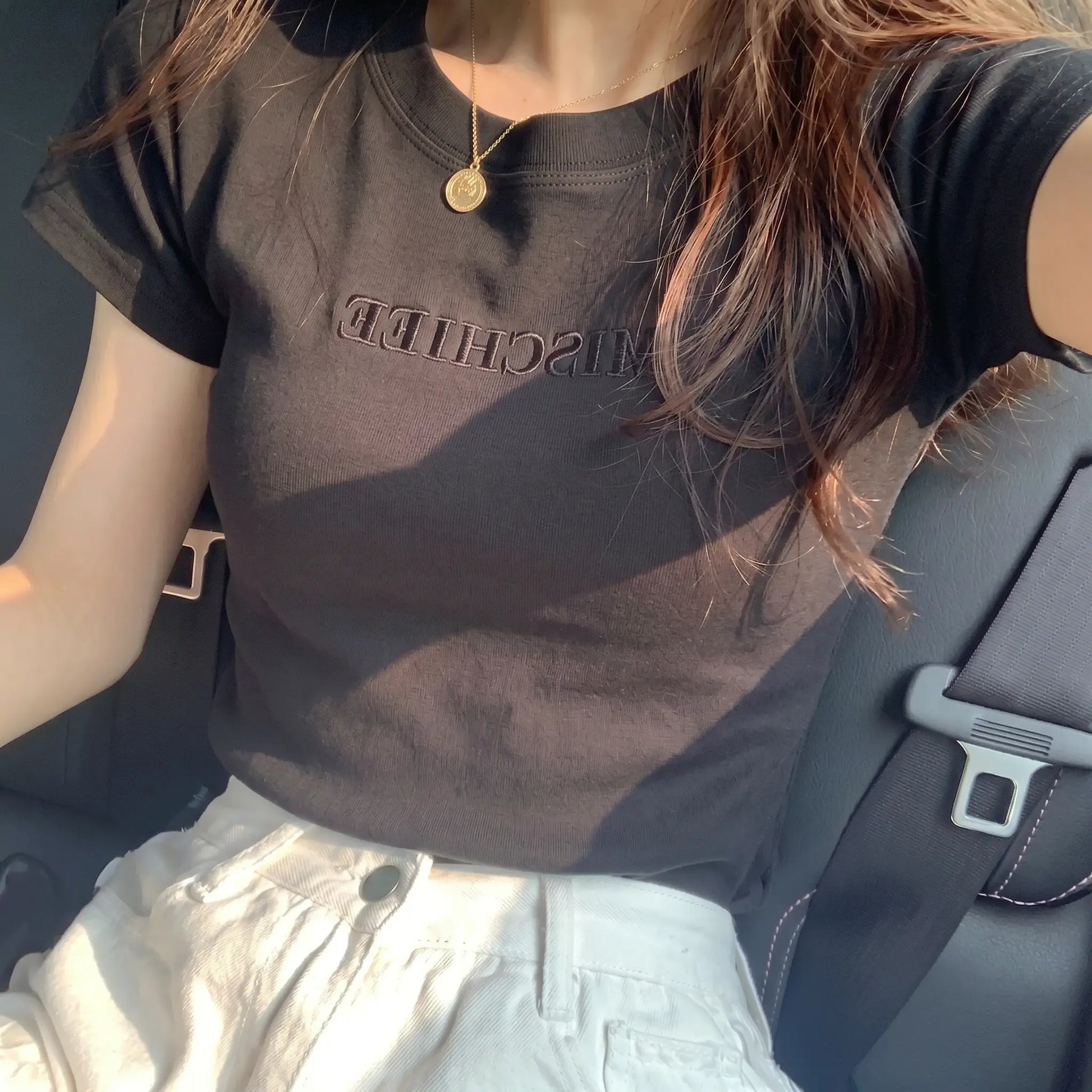 Elegant black slim-fit jersey t-shirt featuring subtle tonal "MISCHIEF" embroidery, worn by a woman in a car, styled with white trousers and a delicate gold pendant necklace.