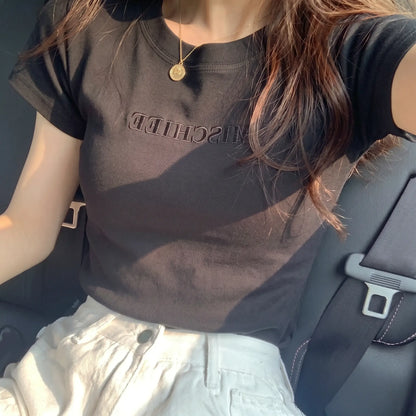 Elegant black slim-fit jersey t-shirt featuring subtle tonal "MISCHIEF" embroidery, worn by a woman in a car, styled with white trousers and a delicate gold pendant necklace.