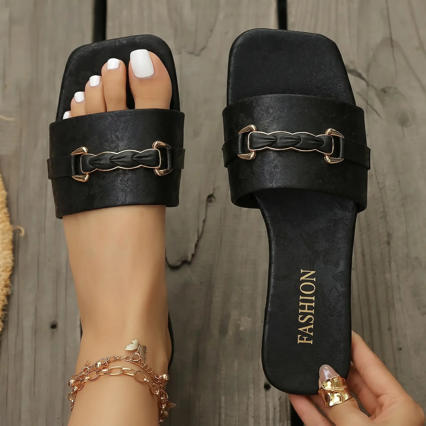 Sophisticated black square-toe slide sandals featuring an embossed floral strap and gold-tone braided buckle, shown on a foot with white pedicure on a wooden surface.