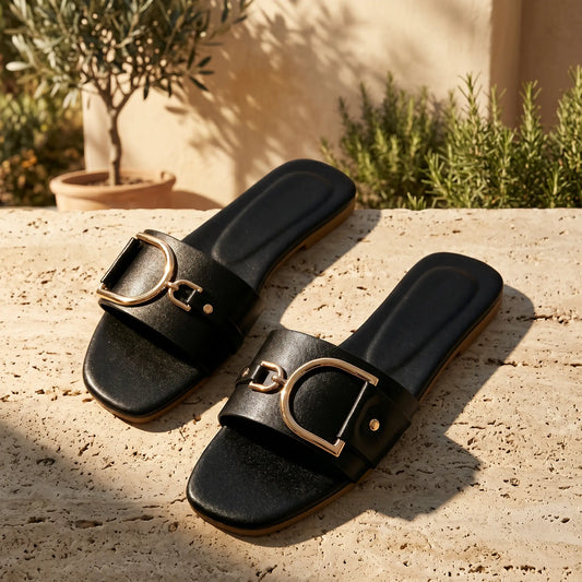 Pair of black square-toe flat slide sandals, featuring wide vegan leather straps with oversized gold-tone horseshoe buckles. Styled on a textured stone surface with natural sunlight and foliage in the background.