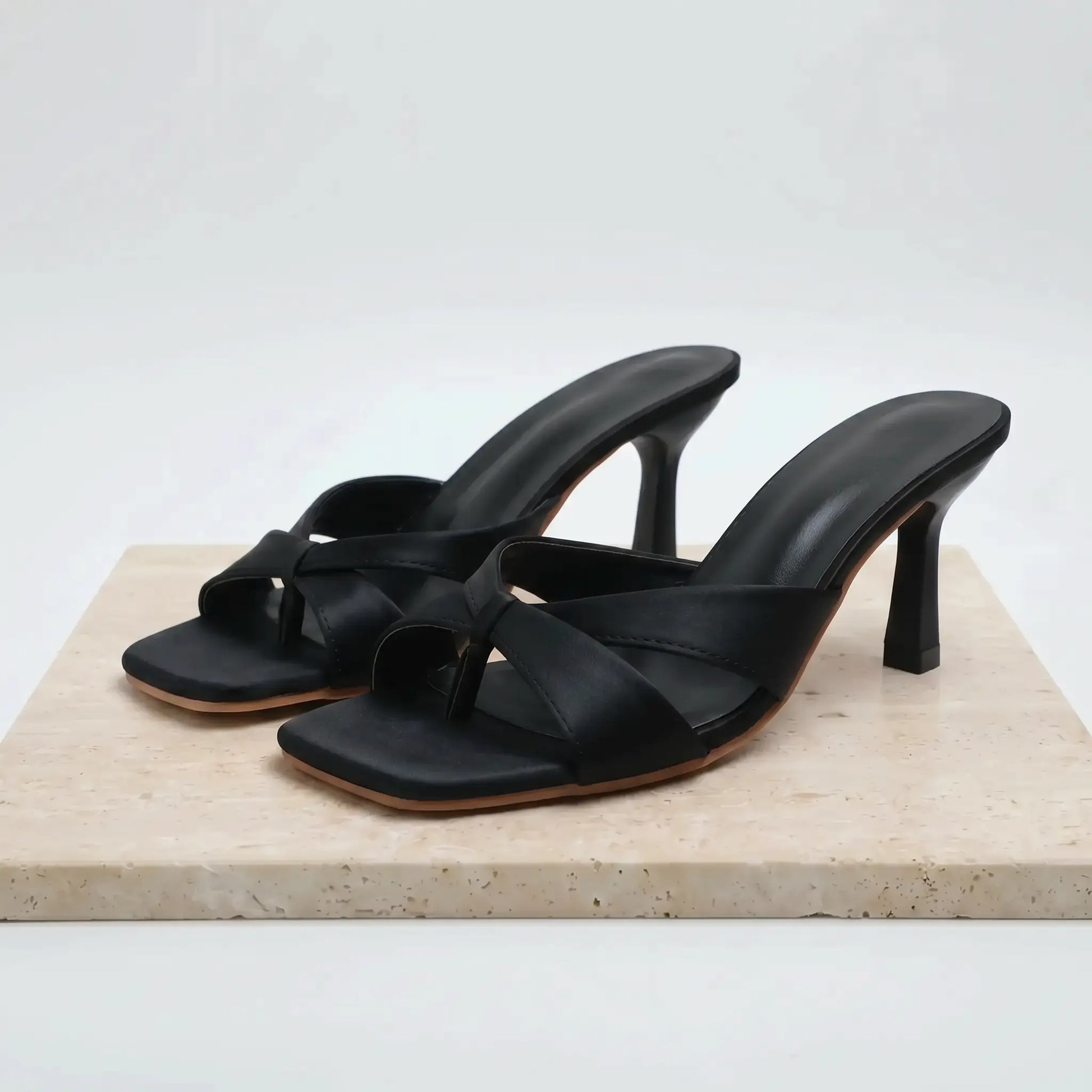 Black PU square toe stiletto mules with a knot detail across the vamp, shown from a three-quarter angle on a marble display.