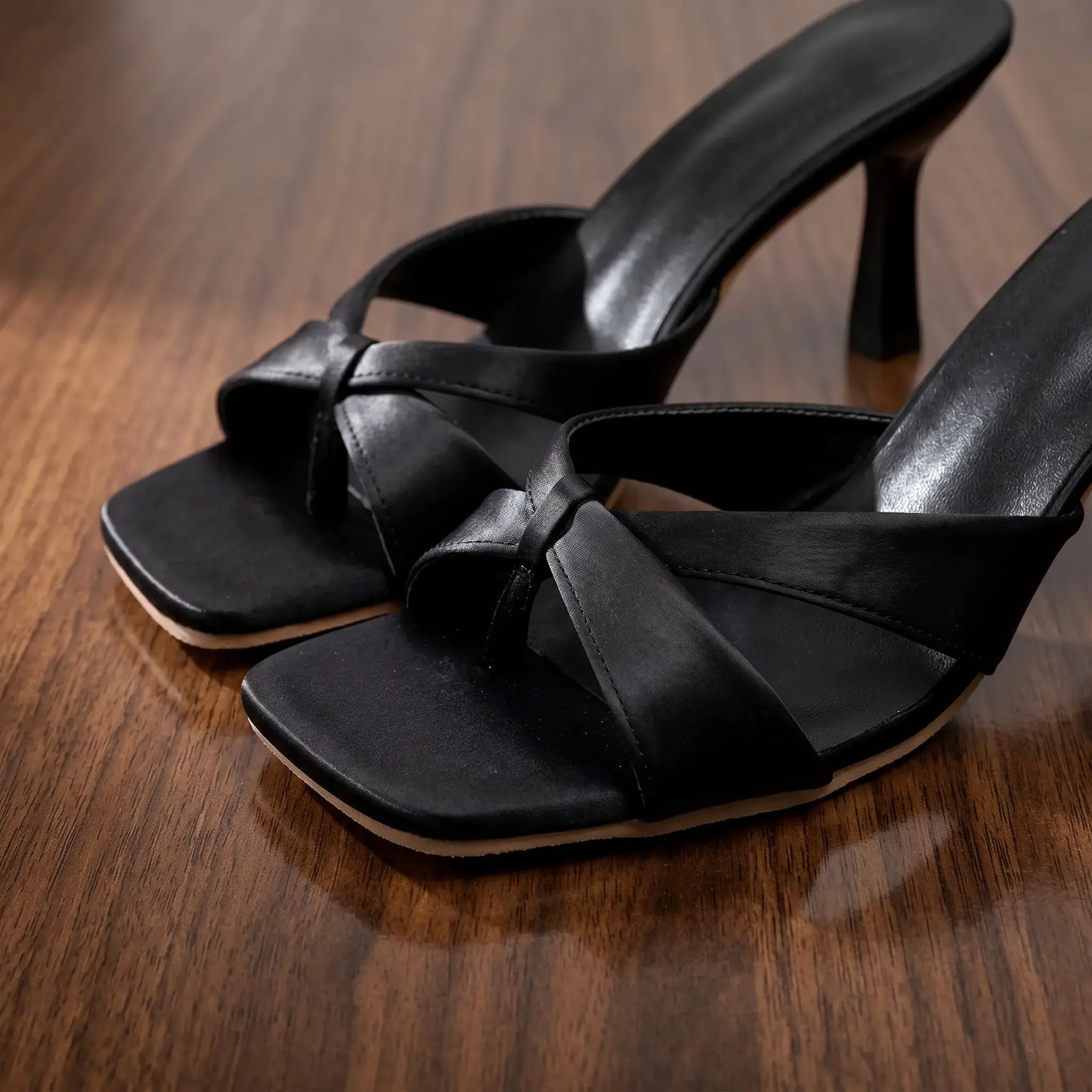 Close-up of black glossy PU square-toe stiletto mules featuring a knot detail across the vamp and a flared stiletto heel, presented on a polished wooden surface.