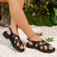 Elegant black strappy flat sandals shown on a woman's feet, detailed with interwoven straps and a gold buckle, set against a natural backdrop of white pebbles and greenery.