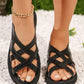 Elegant black strappy flat sandals for women, featuring criss-cross interwoven synthetic leather straps, a flat rubber sole, and a gold-toned ankle bracelet. Displayed on feet against a backdrop of white pebbles and green foliage.
