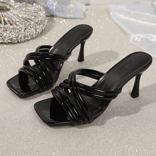 Black high-shine PU strappy heeled mules with a square toe, elegantly crossing over the foot with slender straps. These slip-on sandals feature a flared sculptural heel and a subtle 'FASHION' logo embossed on the insole.