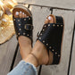 Black studded platform slide sandals with gold buckle, shown on feet with white pedicure on a wooden deck, complementing a straw hat.