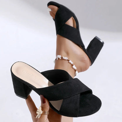 Black suede-like cross-strap block heel mules shown on a model's foot with an anklet, featuring an open toe and a comfortable 7cm block heel.