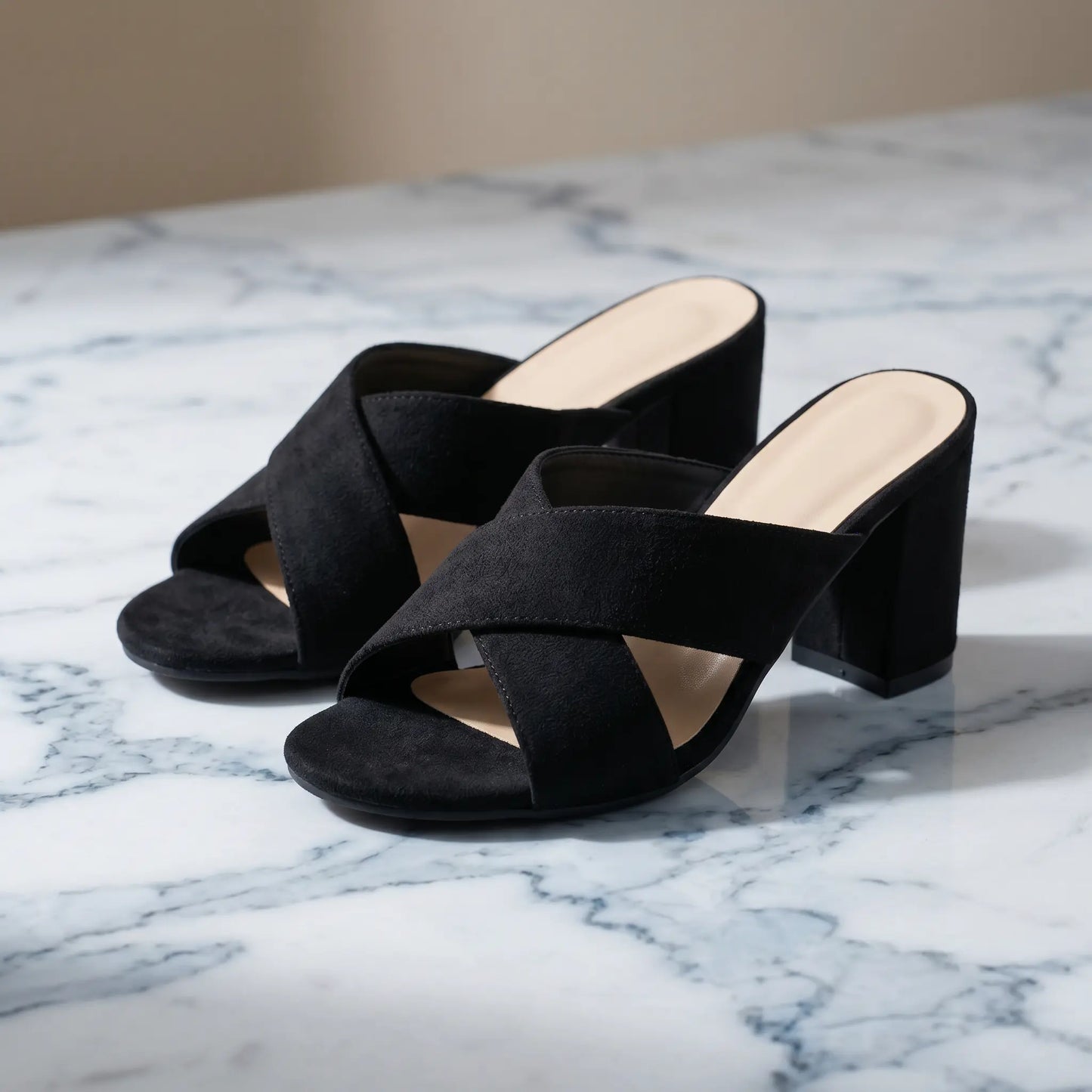 Black suede-like cross-strap block heel mules shown at a three-quarter angle on a marble surface. These open-toe mules feature a comfortable block heel and a sophisticated design.