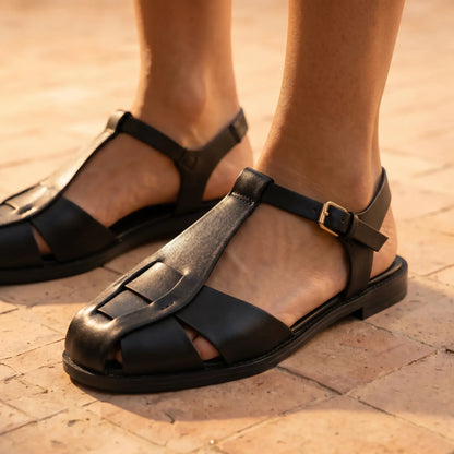 Black Kevroz woven vegan leather fisherman sandals worn on a model's foot, featuring a T-strap, square toe, and gold-tone buckle on a textured light brown tiled surface.