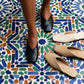 Black vegan leather V-vamp mule flats worn on feet, standing on a vibrant mosaic tile floor, with beige and khaki pairs displayed nearby.