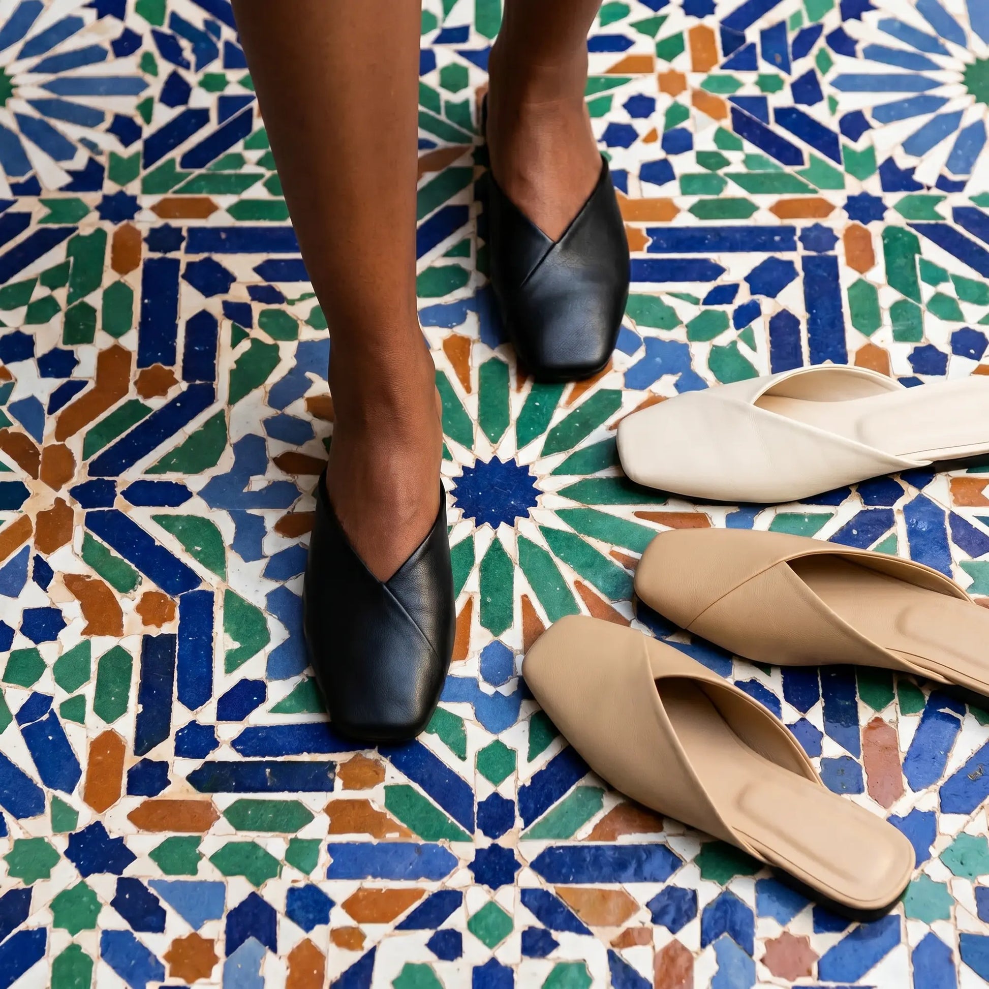 Black vegan leather V-vamp mule flats worn on feet, standing on a vibrant mosaic tile floor, with beige and khaki pairs displayed nearby.