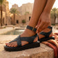 Woman wearing black vegan leather platform Roman sandals with an antique brass buckle, adjusting the ankle strap. The sandals feature a chunky sole and cross-straps with a toe loop, set against a blurred resort background with stone architecture and palm trees.
