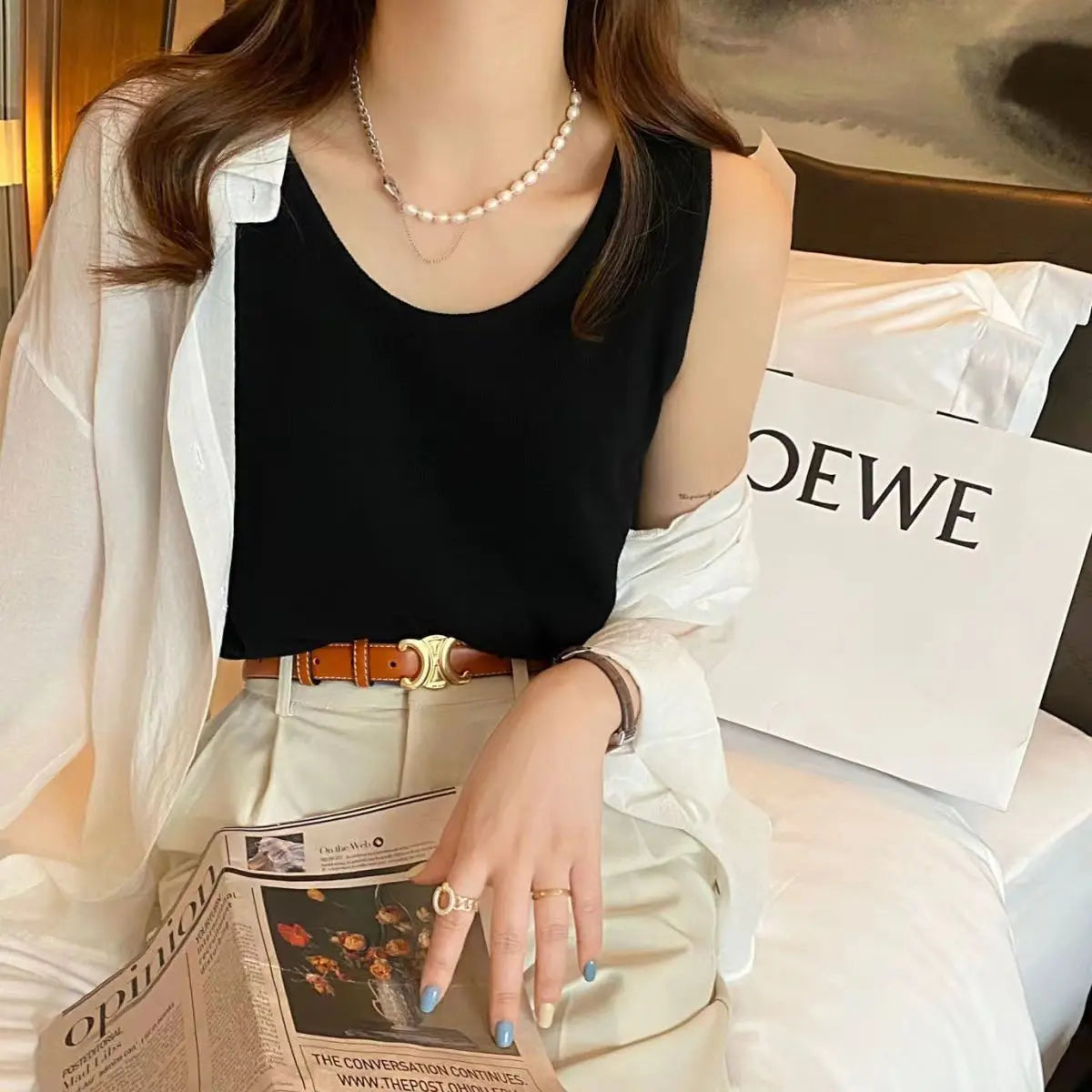 Elegant black waffle knit sleeveless camisole with a scoop neck, worn by a model. The relaxed fit top is layered under a white button-down shirt and styled with light trousers, a brown belt, and a delicate pearl necklace, showcasing its versatile appeal.