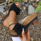 Black Zylaya wedge sandals with a layered floral applique and perforated strap, shown on a foot in an outdoor, stony setting. Features a wood-effect heel and white contrast stitching.
