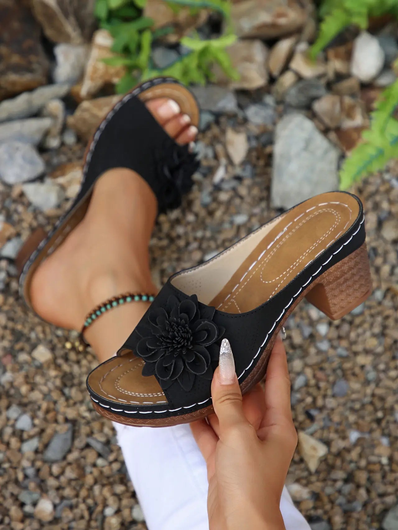Black Zylaya wedge sandals with a layered floral applique and perforated strap, shown on a foot in an outdoor, stony setting. Features a wood-effect heel and white contrast stitching.