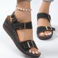 Black faux suede wedge sandals with gold buckle straps and contrast white stitching, shown on a model's feet with a white background. These open-toe sandals feature an adjustable ankle strap and textured wedge heel for comfortable summer style.