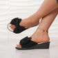 Black fabric wedge slides for women, featuring an oversized bow detail on a single strap and a comfortable wedge heel, shown on a model's feet with a gold anklet.
