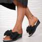 Black Lysa wedge slides with a large fabric bow detail, worn by a model with a black skirt on a textured white background.
