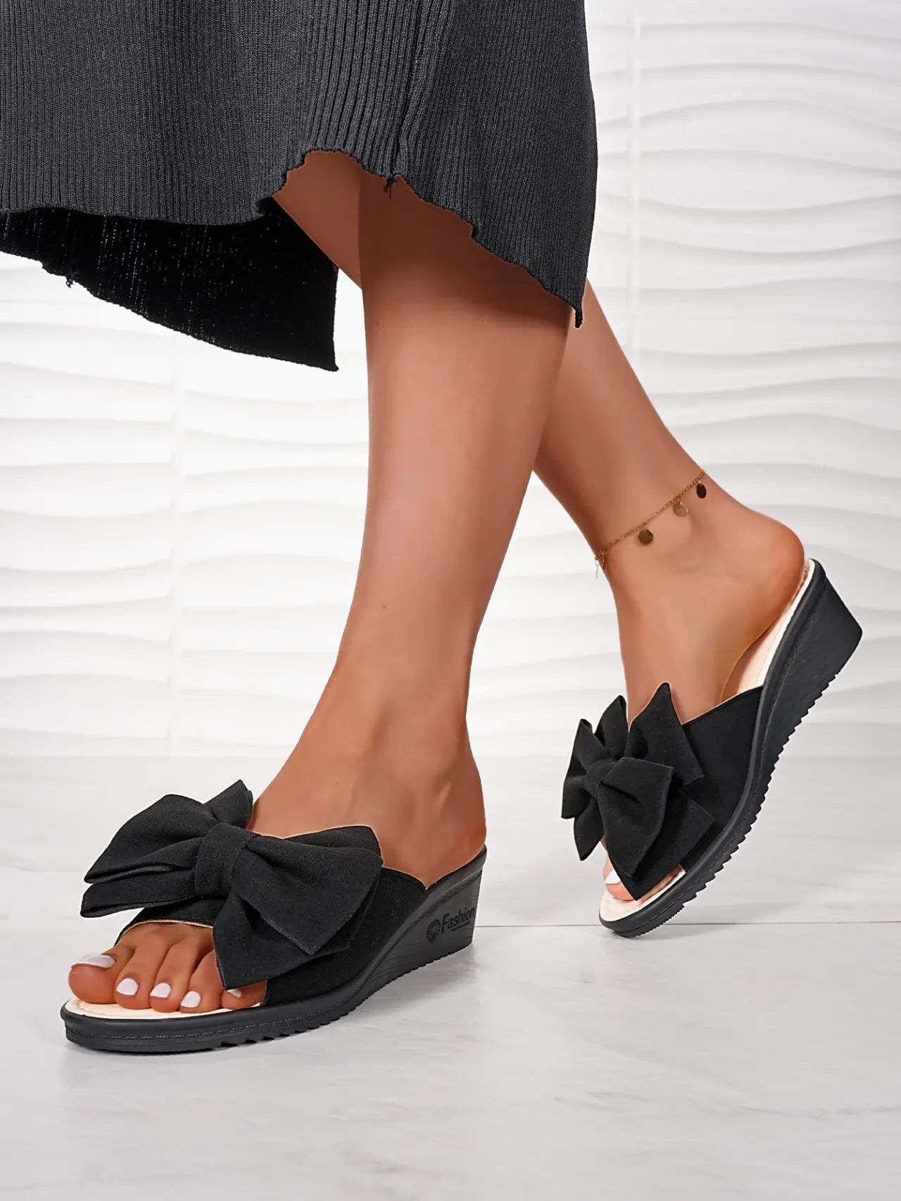 Black Lysa wedge slides with a large fabric bow detail, worn by a model with a black skirt on a textured white background.