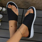 Close-up lifestyle view of a woman's feet wearing black woven thong sandals with a distinctive geometric diamond knit pattern and a crisp white cushioned sole, resting on a wooden deck.