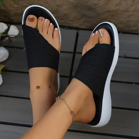 Close-up lifestyle view of a woman's feet wearing black woven thong sandals with a distinctive geometric diamond knit pattern and a crisp white cushioned sole, resting on a wooden deck.