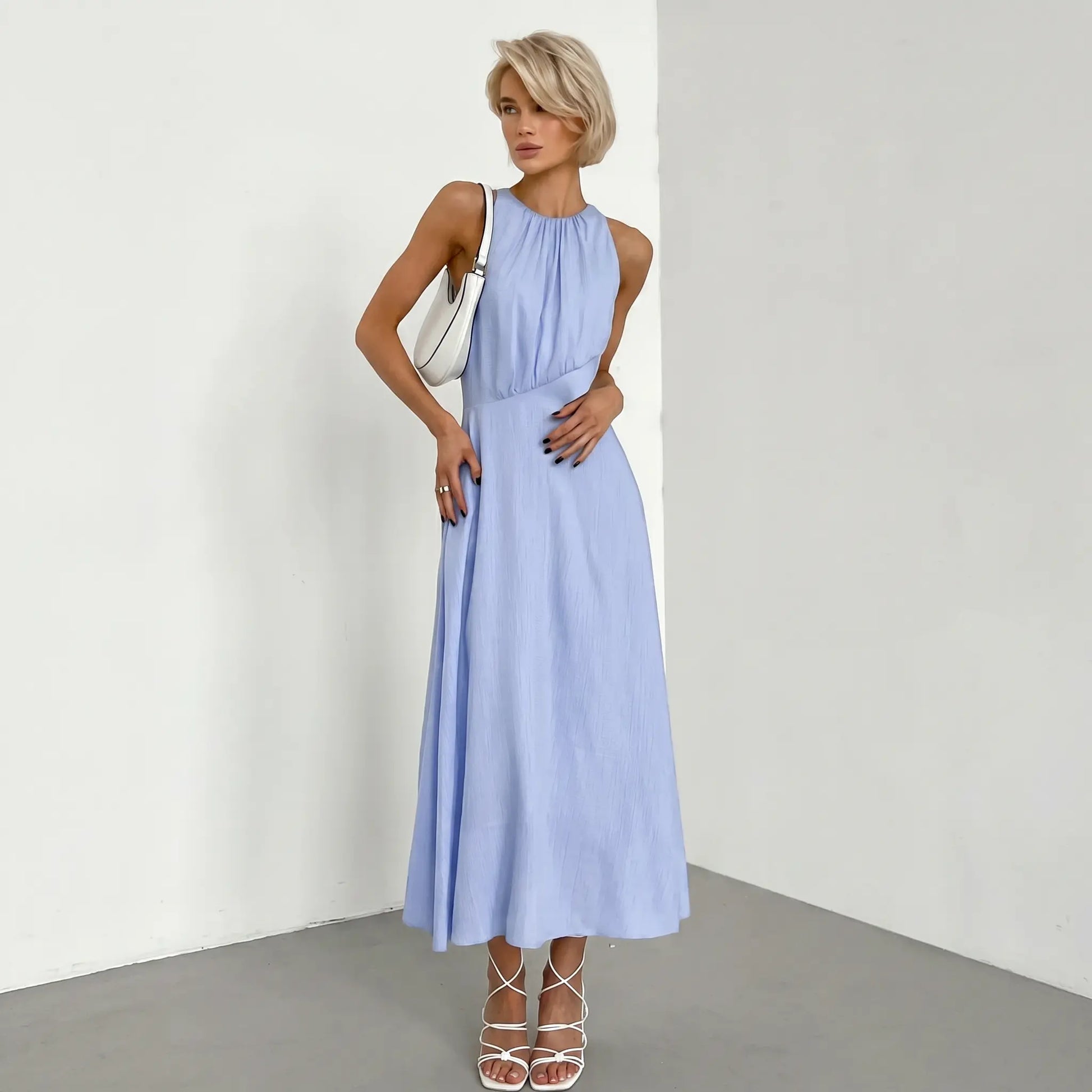 Pale blue sleeveless midi dress with a gathered halter neckline and A-line skirt, styled with a white handbag and white sandals on a model.