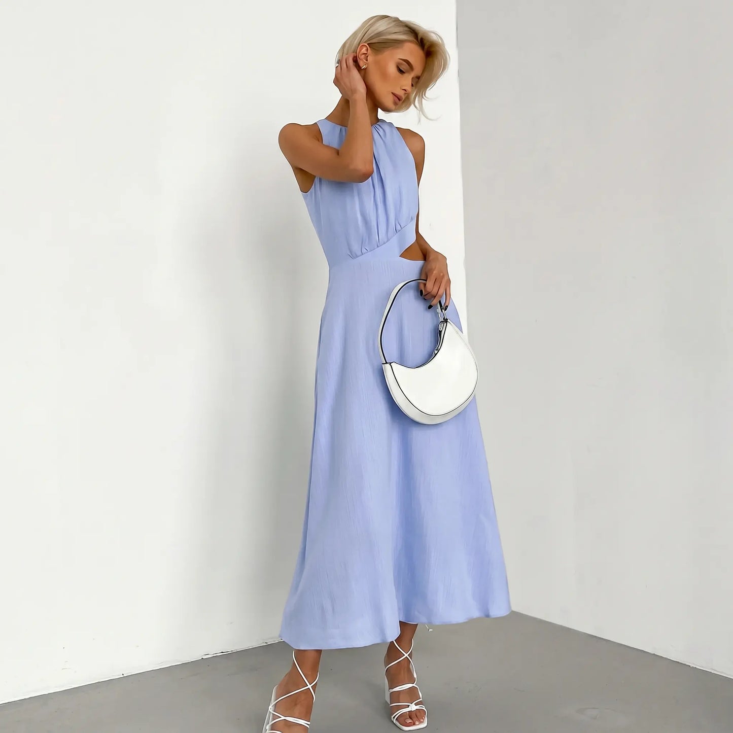 Sleeveless light blue midi dress with gathered halter neckline and side waist cut-outs, worn by a model holding a white handbag, presented against a studio background.
