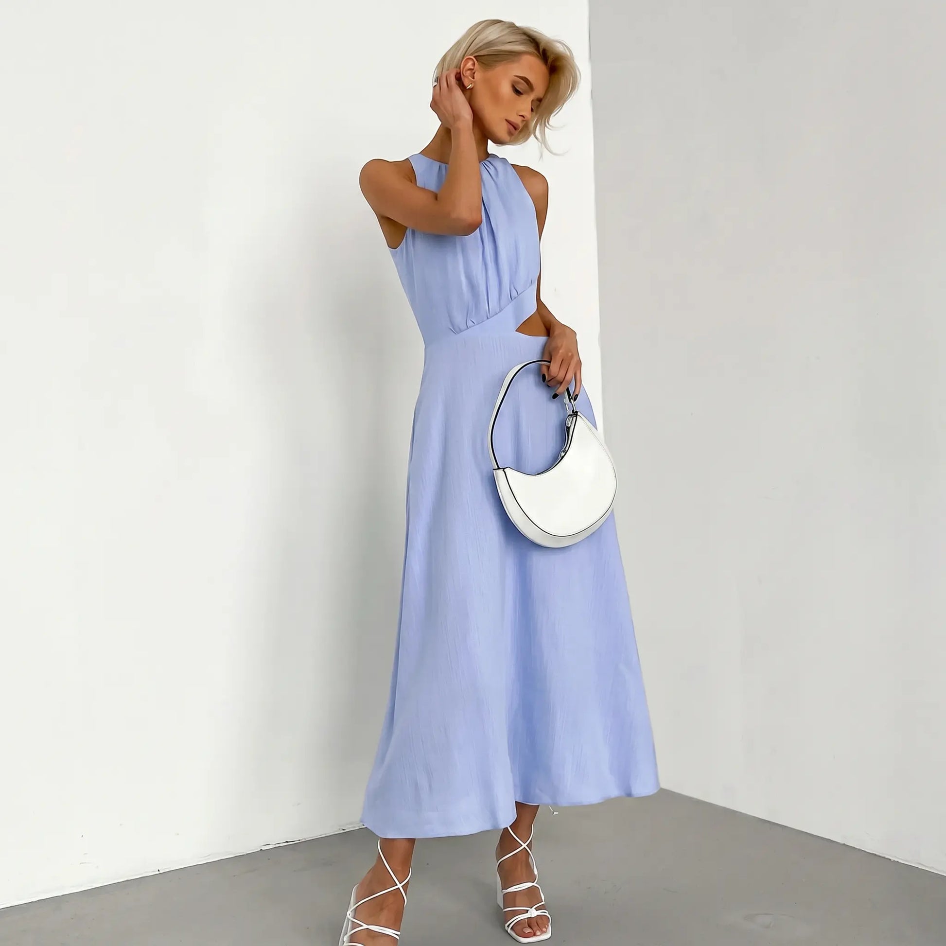 Sleeveless light blue midi dress with gathered halter neckline and side waist cut-outs, worn by a model holding a white handbag, presented against a studio background.