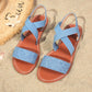 Light blue textured Roman flat sandals with criss-cross elasticated straps and slingback, styled on sand with a straw hat and seashell.