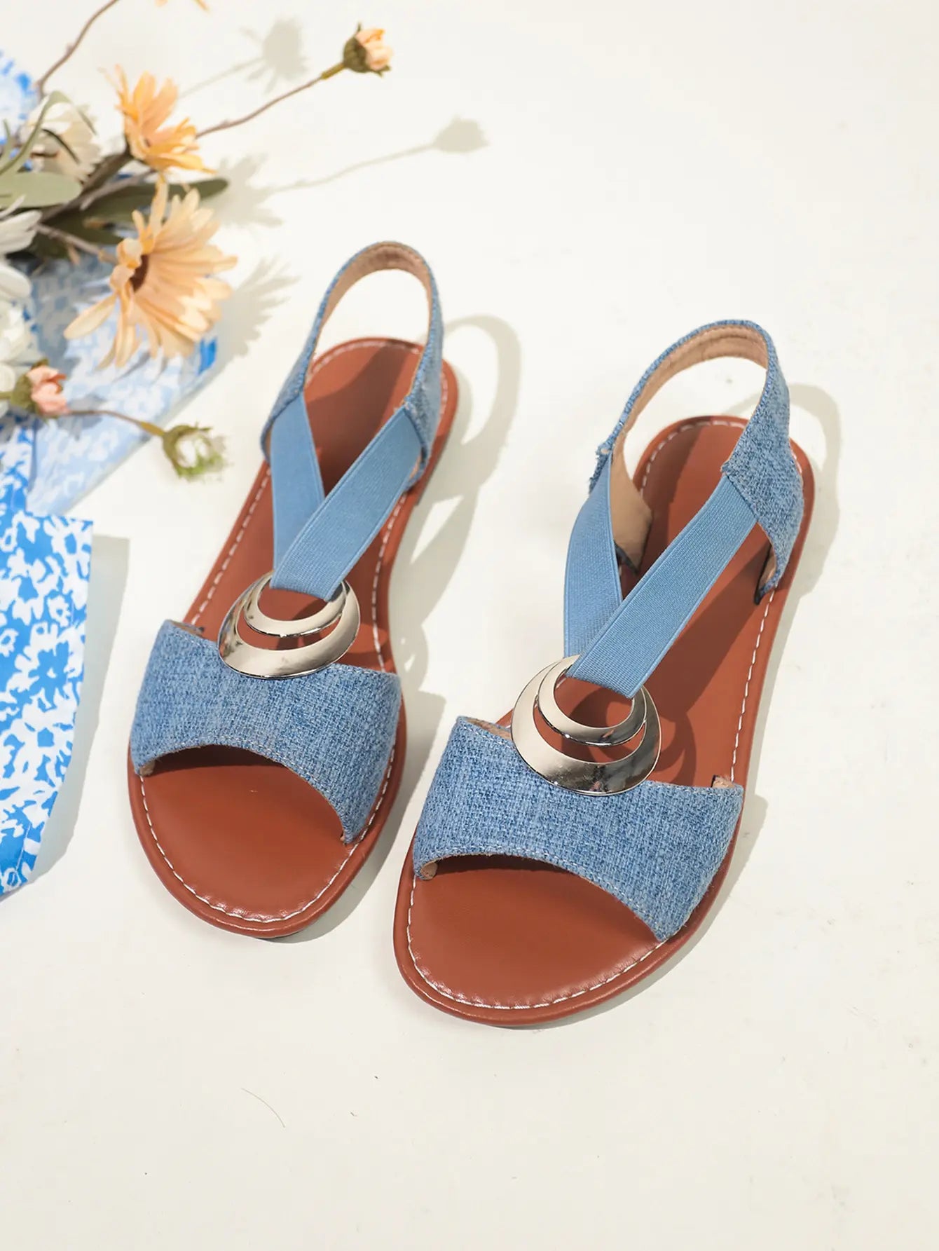 Light blue textured fabric Roman flat sandals with a polished silver-tone metal ring embellishment and elasticated criss-cross slingback straps. The sandals feature a fish mouth peep-toe design and a flat brown footbed with a black rubber sole.