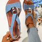 Close-up of light blue textured Roman flat sandals on a foot, featuring a polished silver-tone metal ring embellishment on the vamp strap and crossed elasticated slingback straps.