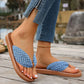 Woman's feet wearing blue woven thong strap flat sandals with a tan sole, paired with blue jeans and a gold anklet, shown from a side view on a wooden deck.
