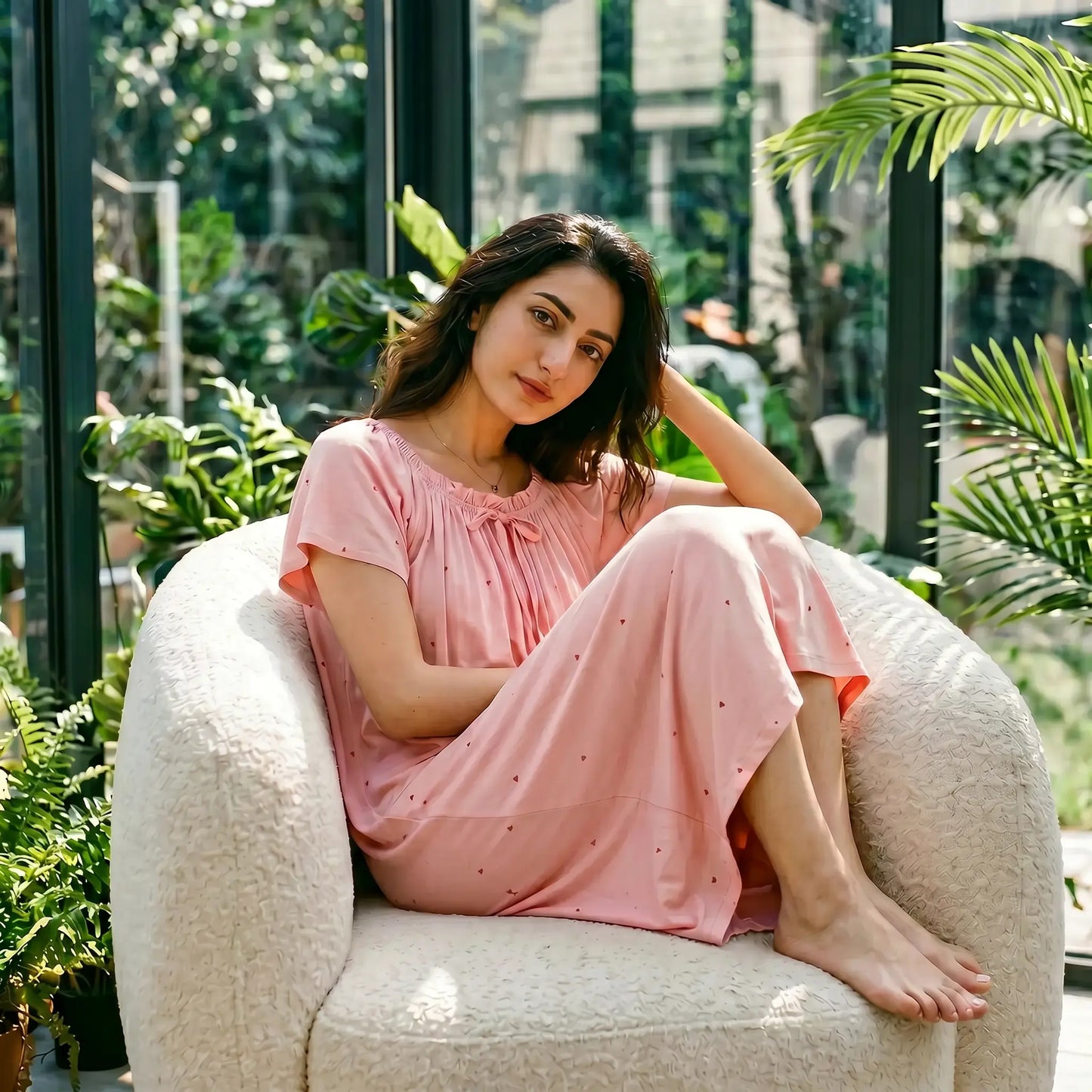 Female model in a Blush Pink Zevryn Modal Romantic Lounge Dress, featuring gathered scoop neck, self-tie bow, and flutter sleeves, relaxing in a sunlit, plant-filled conservatory.