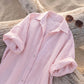 Close-up of an exquisite blush rose pink linen-blend maxi shirt dress, featuring a classic collar, elegant pearl-toned buttons, and casually rolled-up long sleeves, laid flat on a weathered wooden log with soft sand in the background.