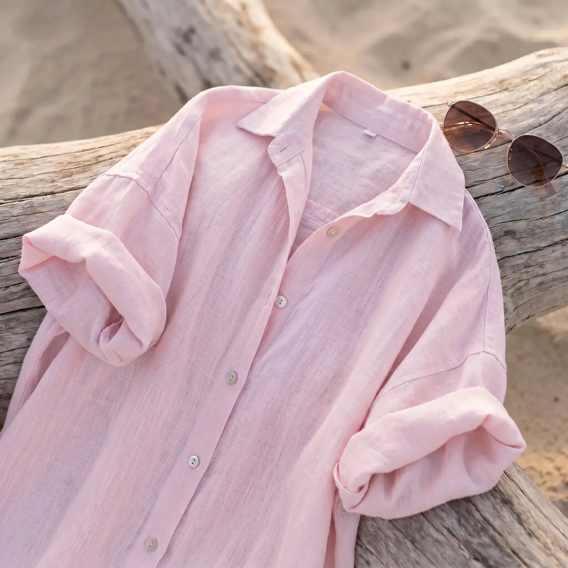 Close-up of an exquisite blush rose pink linen-blend maxi shirt dress, featuring a classic collar, elegant pearl-toned buttons, and casually rolled-up long sleeves, laid flat on a weathered wooden log with soft sand in the background.
