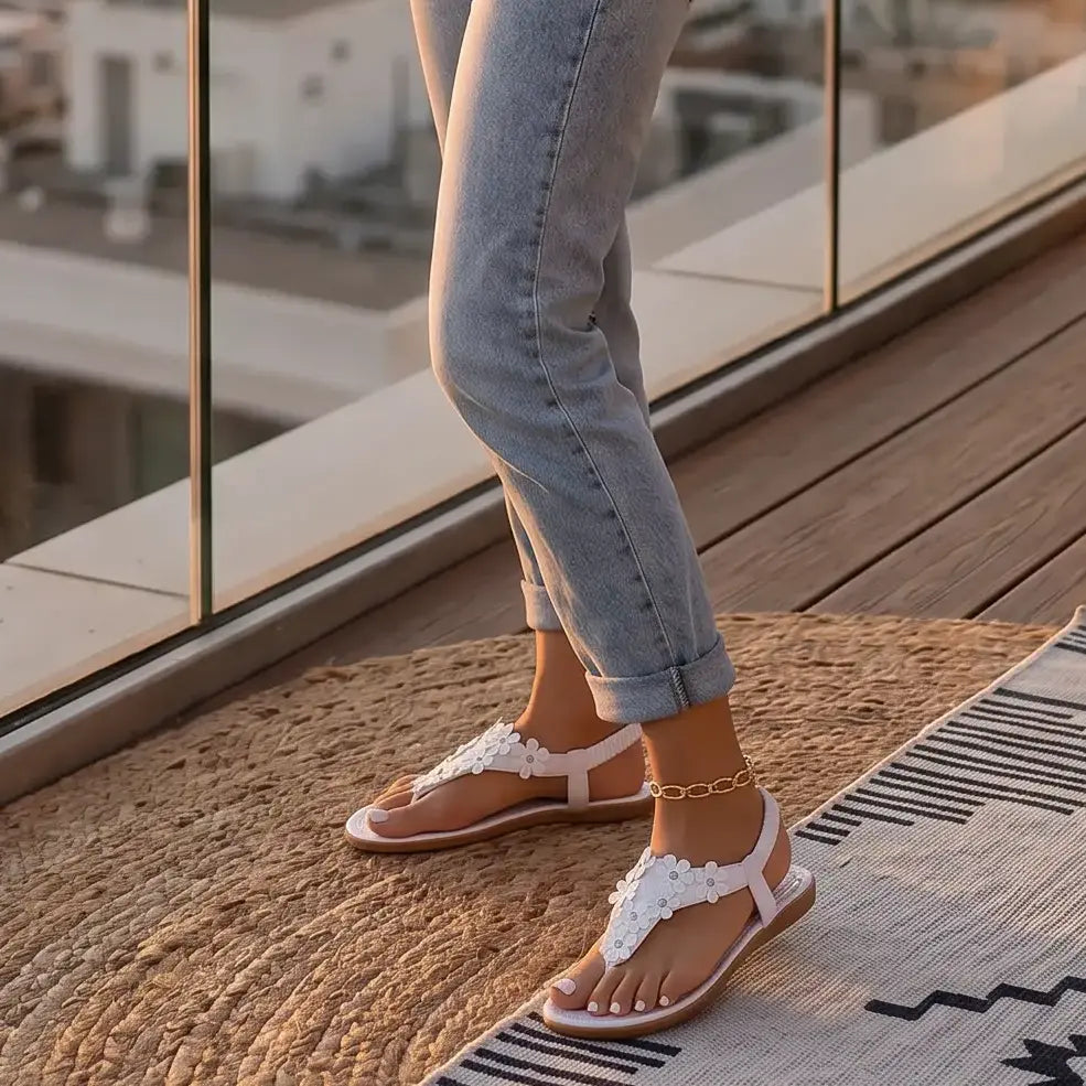 Woman wearing white bohemian flat t-strap sandals with 3D floral appliques and elastic slingback, standing on a rooftop terrace at sunset.