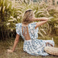 Back view of a woman wearing a bohemian midi dress with a blue floral print, featuring an open back with a tie closure and ruffled cap sleeves, sitting outdoors in tall grass.