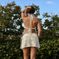 Back view of a woman wearing a sheer off-white floral lace halter crop top and mini skirt set in a sunny outdoor setting.