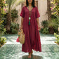 Elegant woman in a flowing Bordeaux Crimson tiered maxi dress with a deep V-neck and crinkled gauze fabric, walking barefoot in a vibrant tropical garden.