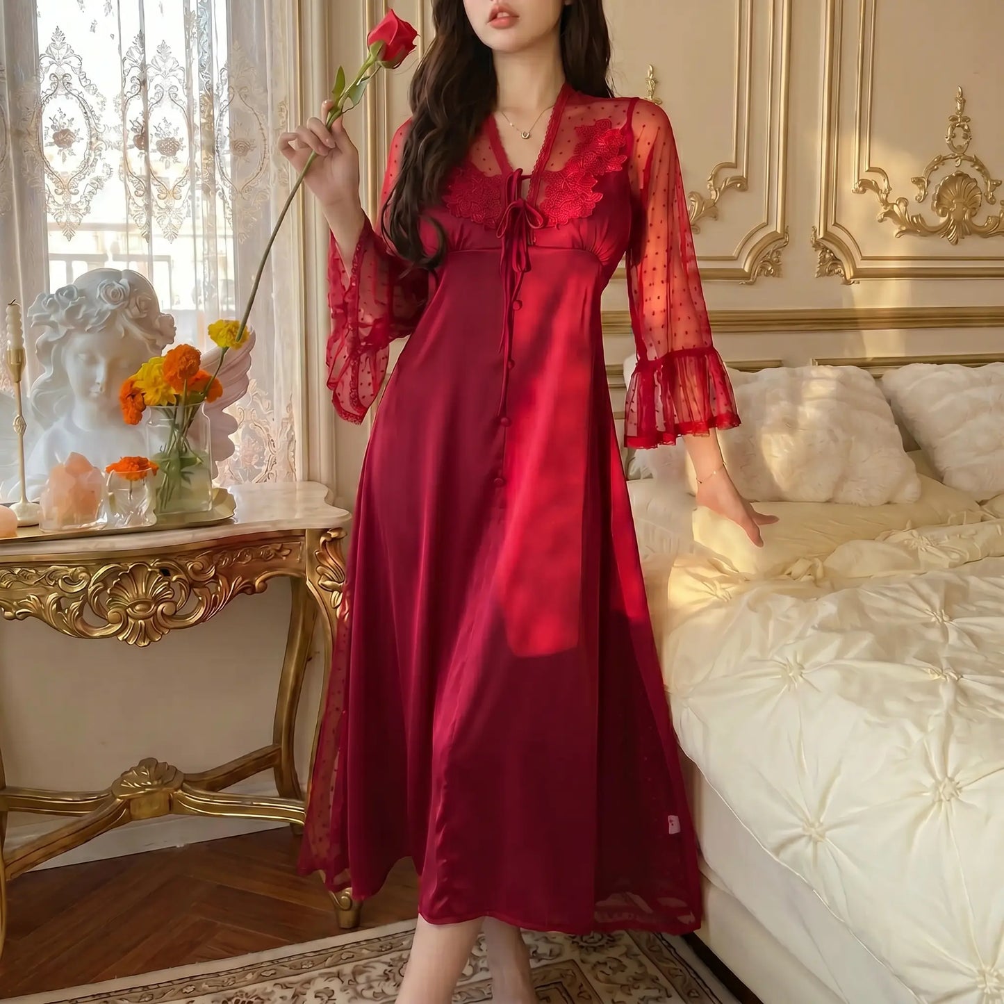 Woman in a Bordeaux Red Sylven long satin nightgown with intricate floral lace V-neck, sheer polka dot bell sleeves, and front button details, holding a red rose in an elegant bedroom setting.
