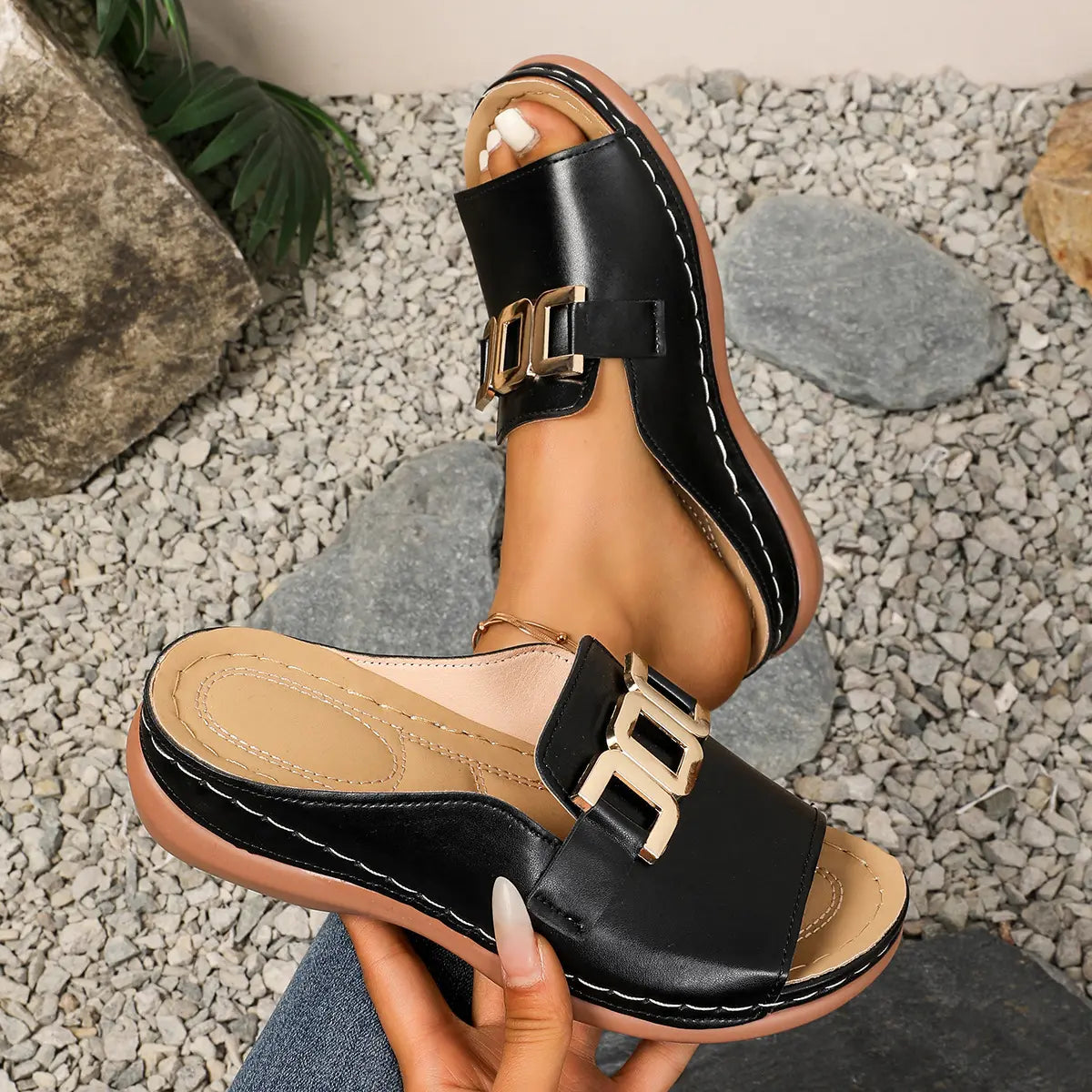 Black Brevix women's platform slide sandals featuring a decorative gold-tone metallic buckle and comfortable contoured footbed, shown worn on a foot against a pebble and natural stone background.