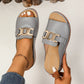 Grey Brevix platform slide sandals with a gold interlocking buckle detail, shown on a woman's foot with a white pedicure and delicate ankle bracelet.