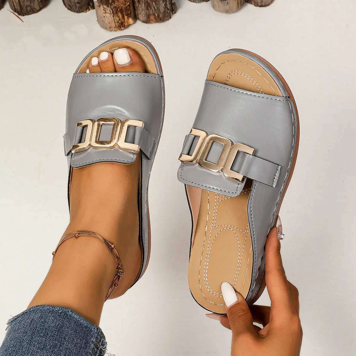 Grey Brevix platform slide sandals with a gold interlocking buckle detail, shown on a woman's foot with a white pedicure and delicate ankle bracelet.