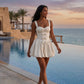 Model walks beside an infinity pool at sunset, wearing a Brevon buttercream yellow bow-detail mini dress with a structured corset bodice and voluminous puff skirt.