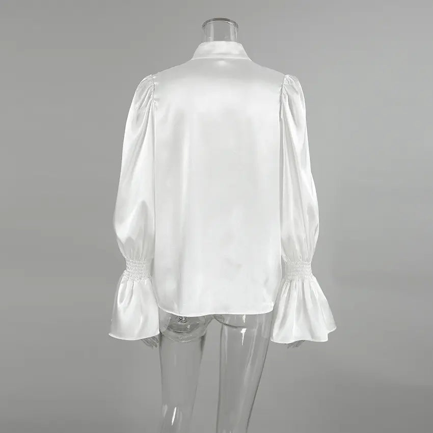 Back view of a white lustrous satin blouse featuring a high stand collar, dramatic ruffled front, and voluminous trumpet sleeves with smocked elasticated cuffs.