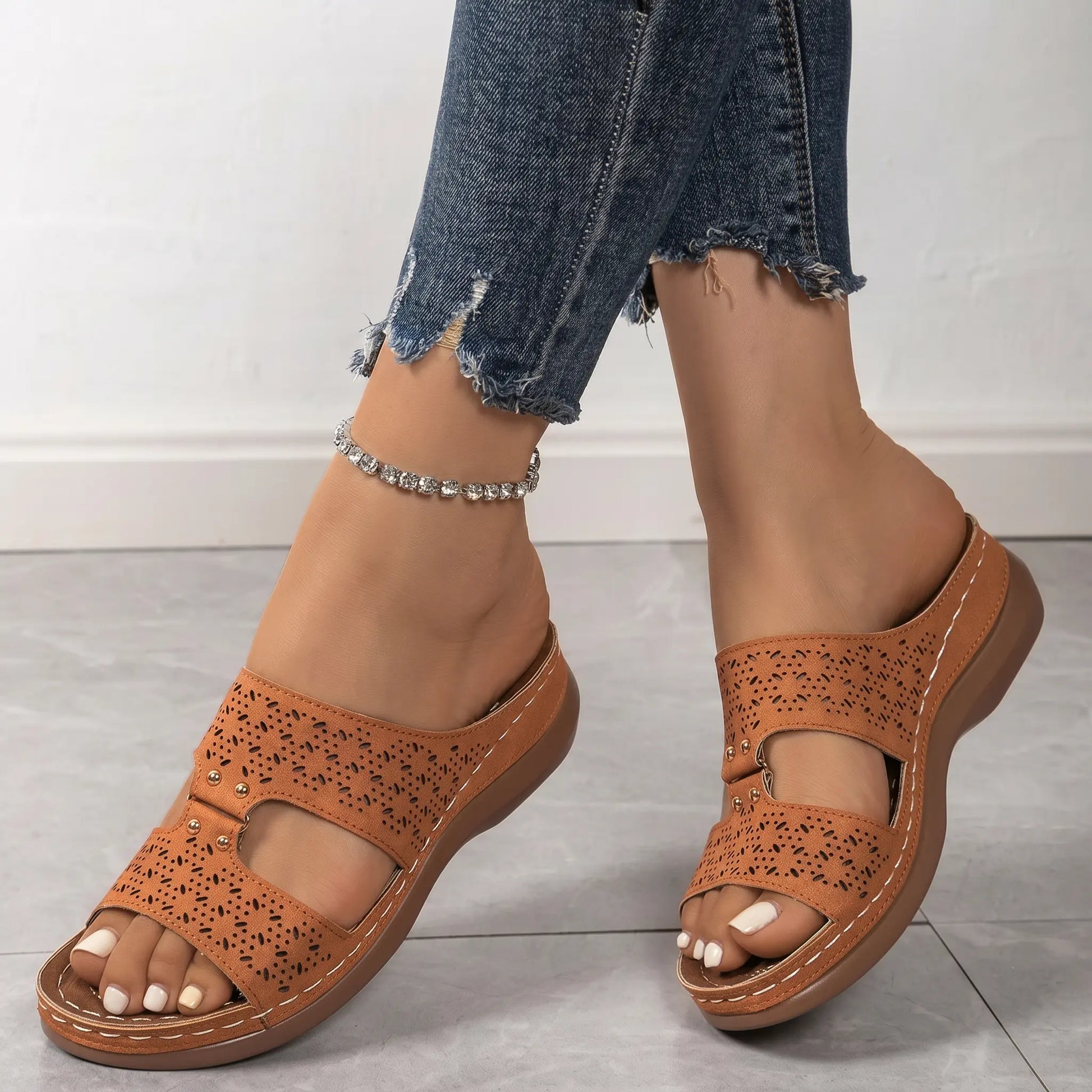 Woman's feet wearing tan Briska laser-cut slip-on wedge sandals with distressed blue jeans and a silver anklet, showcasing intricate detailing and comfort.