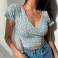 Model wears a light blue Brivan cropped faux-wrap V-neck top with delicate white floral print and short cap sleeves, styled with high-waisted denim jeans and a gold pendant necklace.
