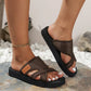 Woman's feet wearing brown textured fabric criss-cross platform slides with an embossed 'FASHION' tab, styled on natural stones and pebbles.