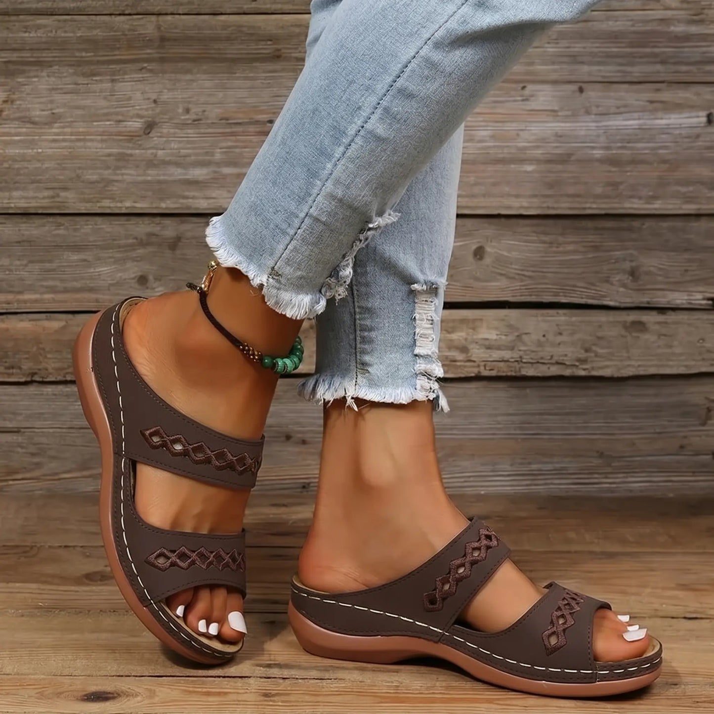 A woman's feet wearing dark brown slip-on slide sandals with intricate geometric embroidered straps and white contrast stitching. She wears distressed denim jeans, showcasing a casual summer look.