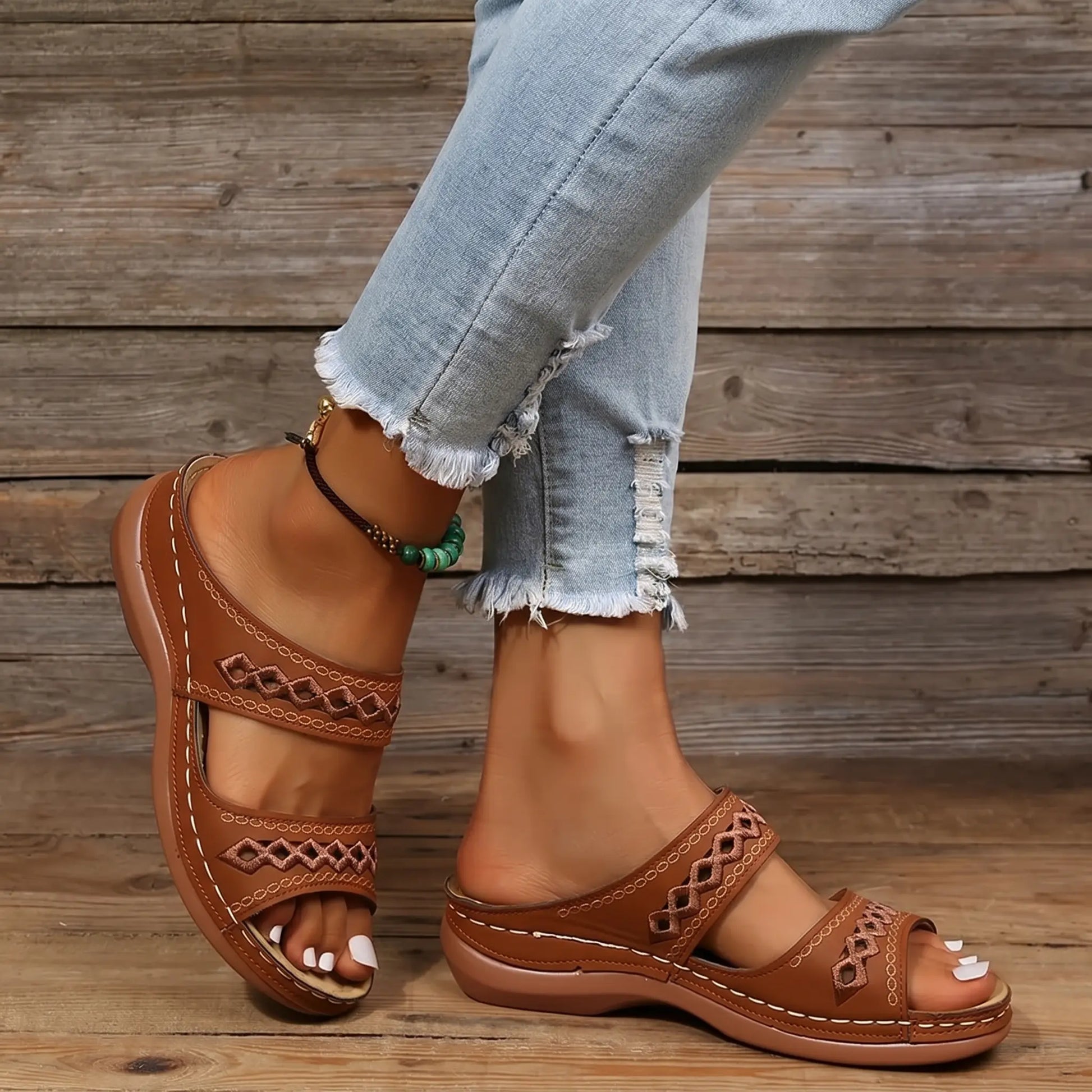 Women's warm chestnut PU leather slide sandals featuring double straps with intricate geometric embroidery, worn with light-wash distressed denim jeans.
